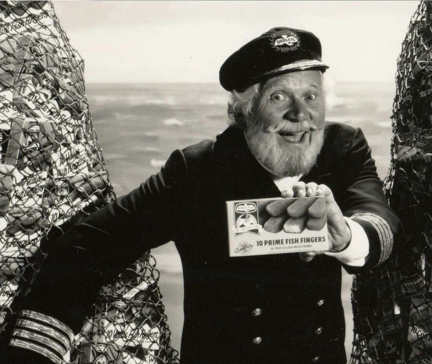 Black-and-white advertising photograph of Captain Birds Eye holding a box of fish fingers