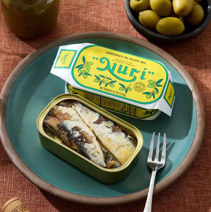 An open tin of NURI sardines in olive oil beside its paper wrapper on a green plate